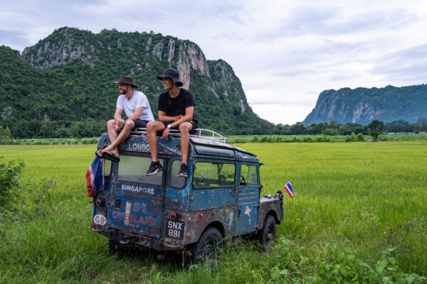 Two men sit on an old blue coloured Land Rover, with their legs dangling over the edge. The car in sitting in the middle of a grass field with mountains in the background.